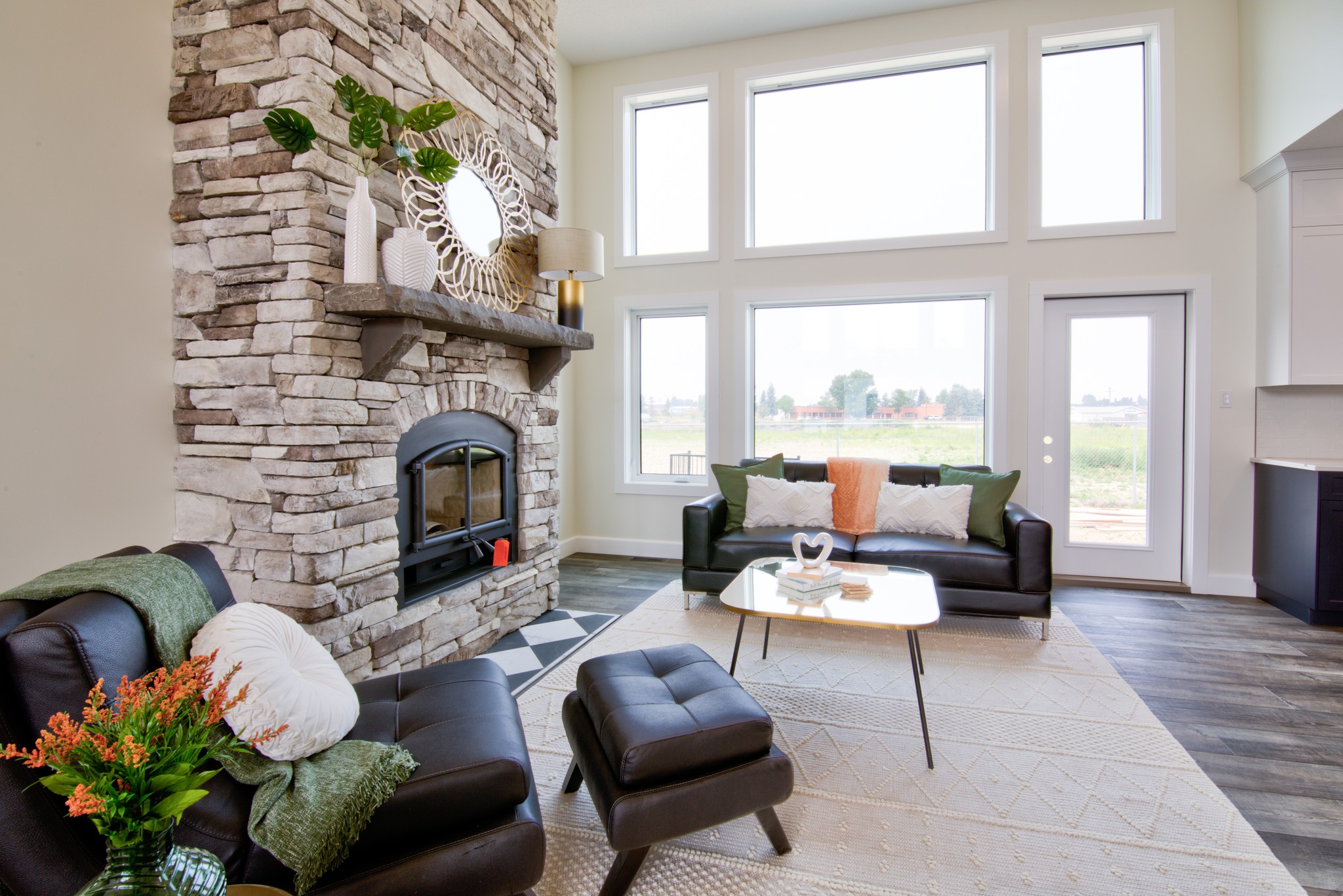 A cozy living room with large windows, a stone fireplace, and a dark leather sofa set. Bright and airy, with modern decor and neutral tones.