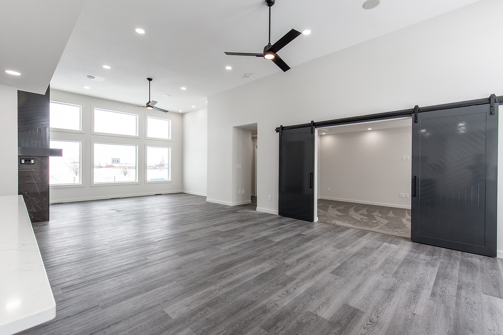 Spacious modern living area with dark wood flooring, white walls, and large windows. Features black sliding barn doors and ceiling fans. Bright, open ambiance.