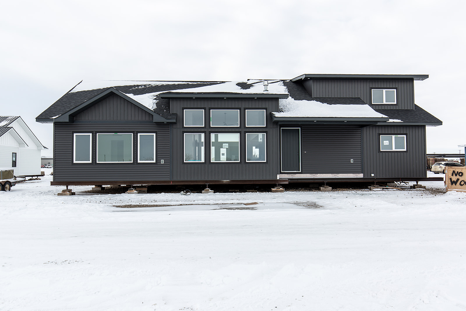 Modern gray modular home on a snowy lot, elevated on blocks. Large windows and snowy roof contrast with overcast sky, conveying a serene winter feel.