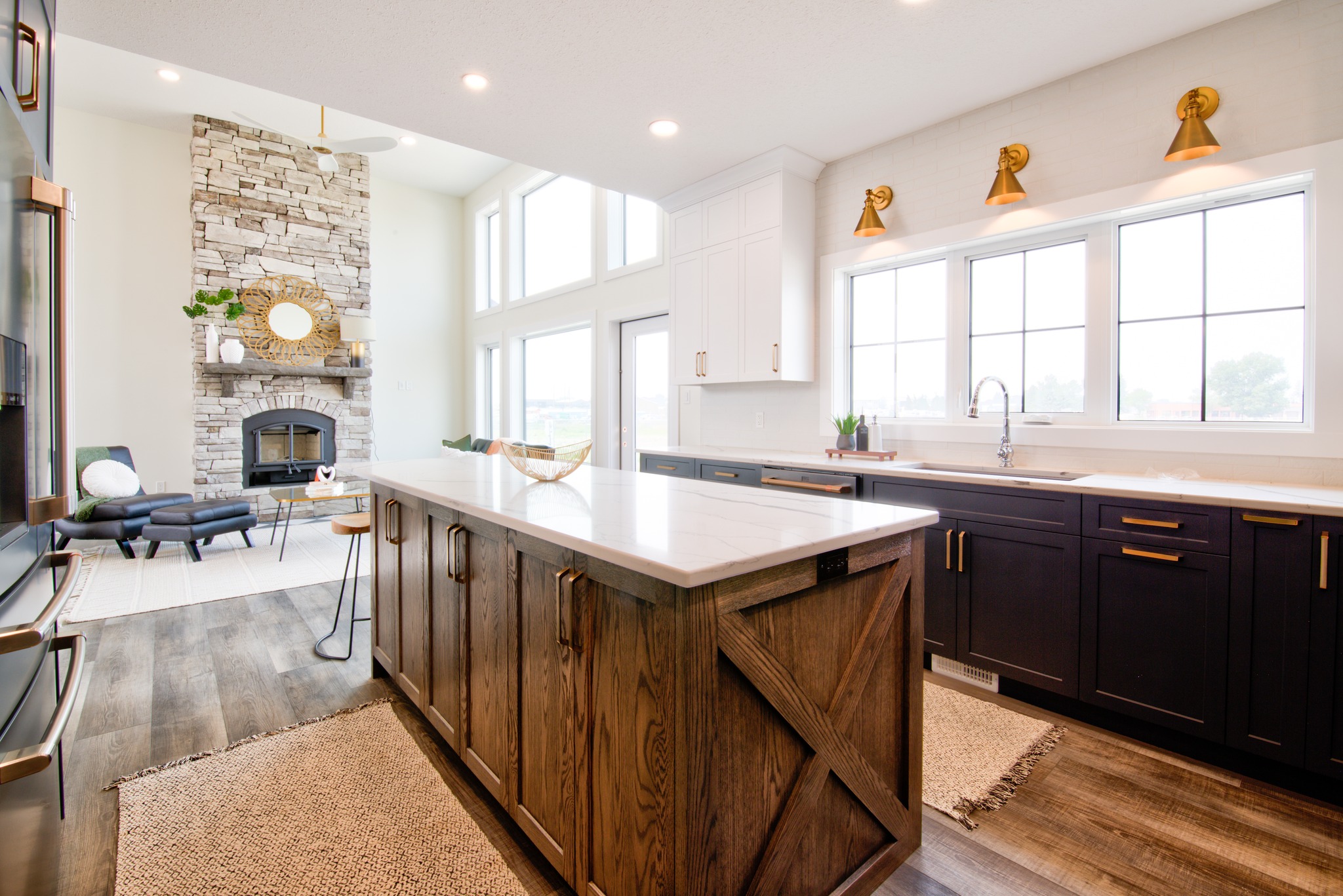 Bright kitchen with a large island featuring wood cabinets and a marble countertop. Floor-to-ceiling windows illuminate the space. A stone fireplace and cozy seating area add warmth and sophistication.
