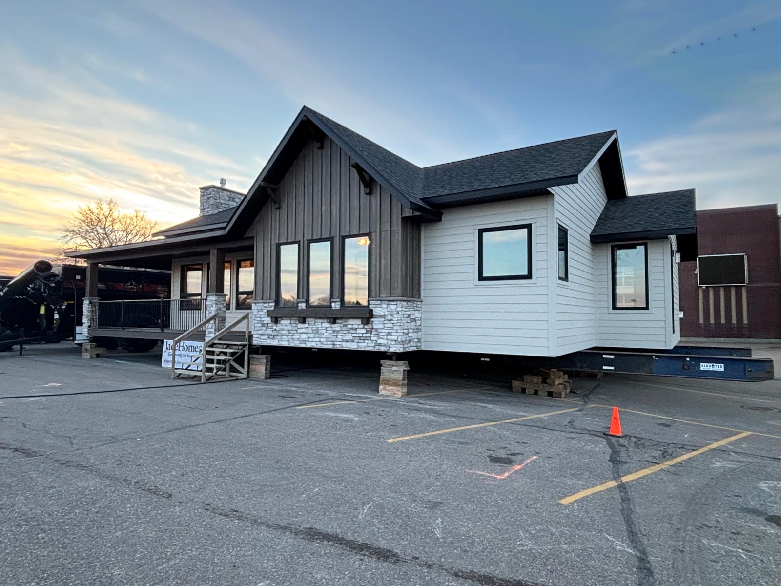 A modern prefabricated home sits elevated on a platform in a parking lot, surrounded by a scenic sunset. The house features wood paneling, large windows, and a stone chimney.