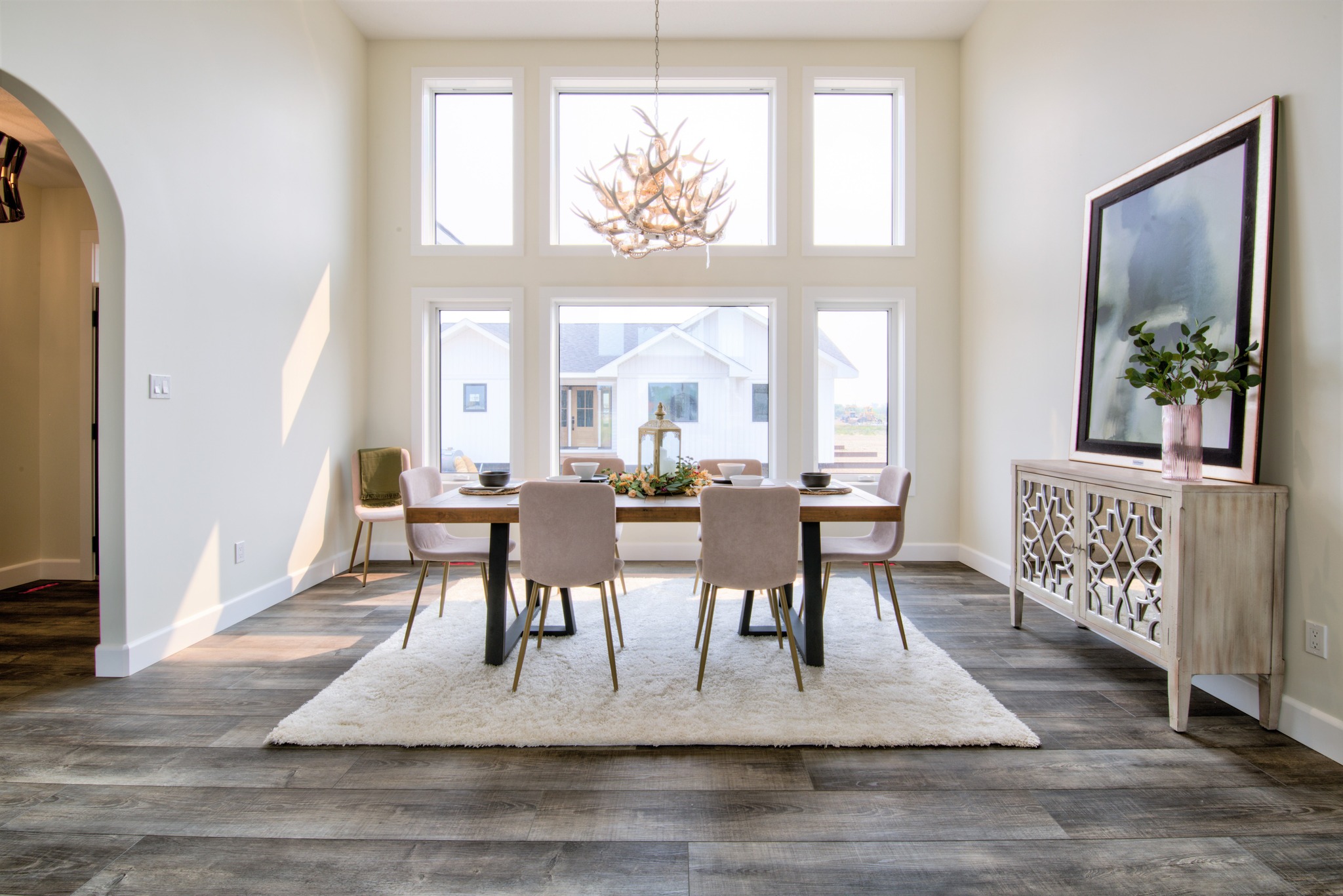 Bright dining room with large windows, wooden floor, elegant table set on a white rug, and a decorative sideboard with a plant. Airy and inviting.