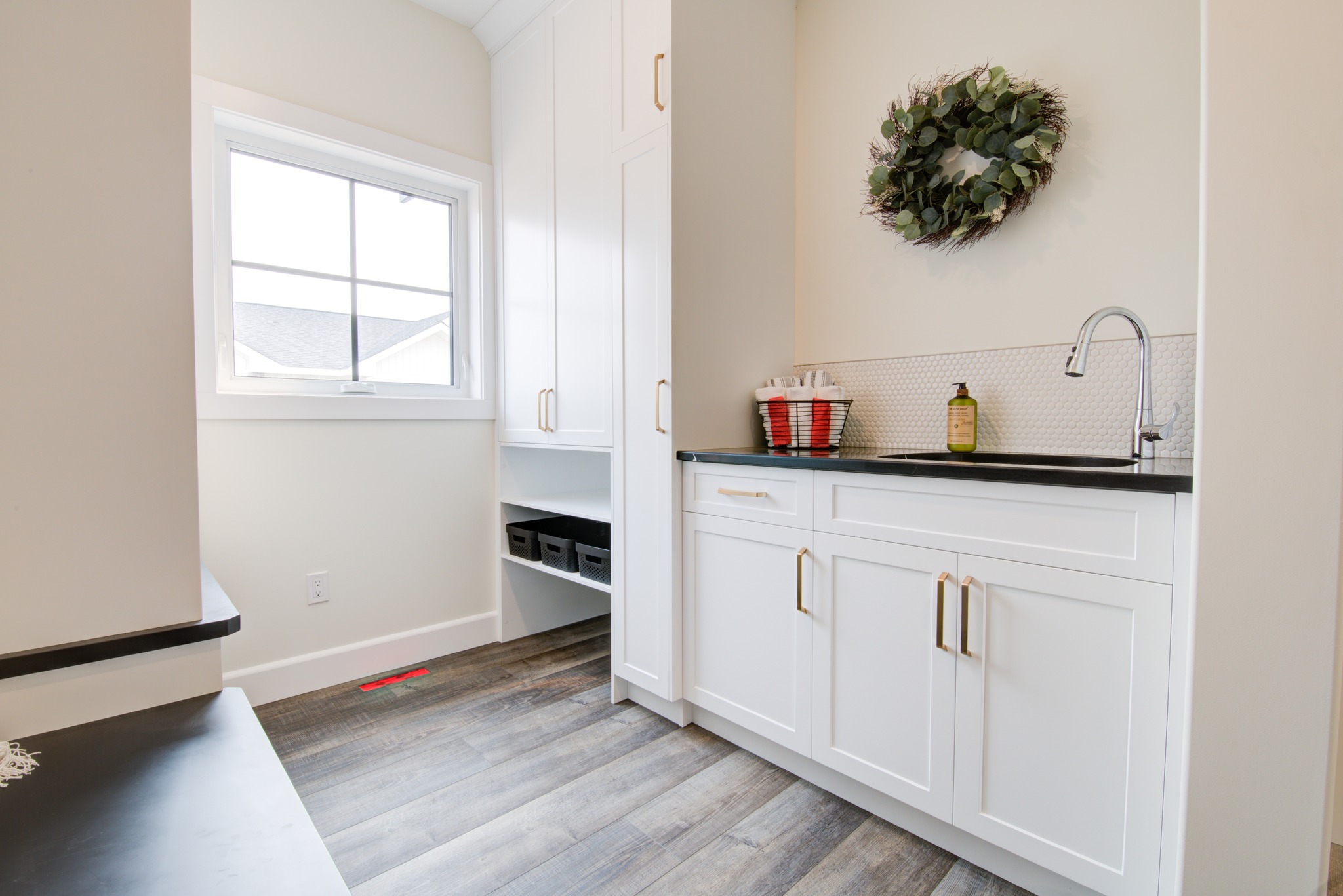 Bright laundry room with white cabinets, dark countertops, and a farmhouse sink. A wreath decorates the wall, creating a fresh, welcoming atmosphere.
