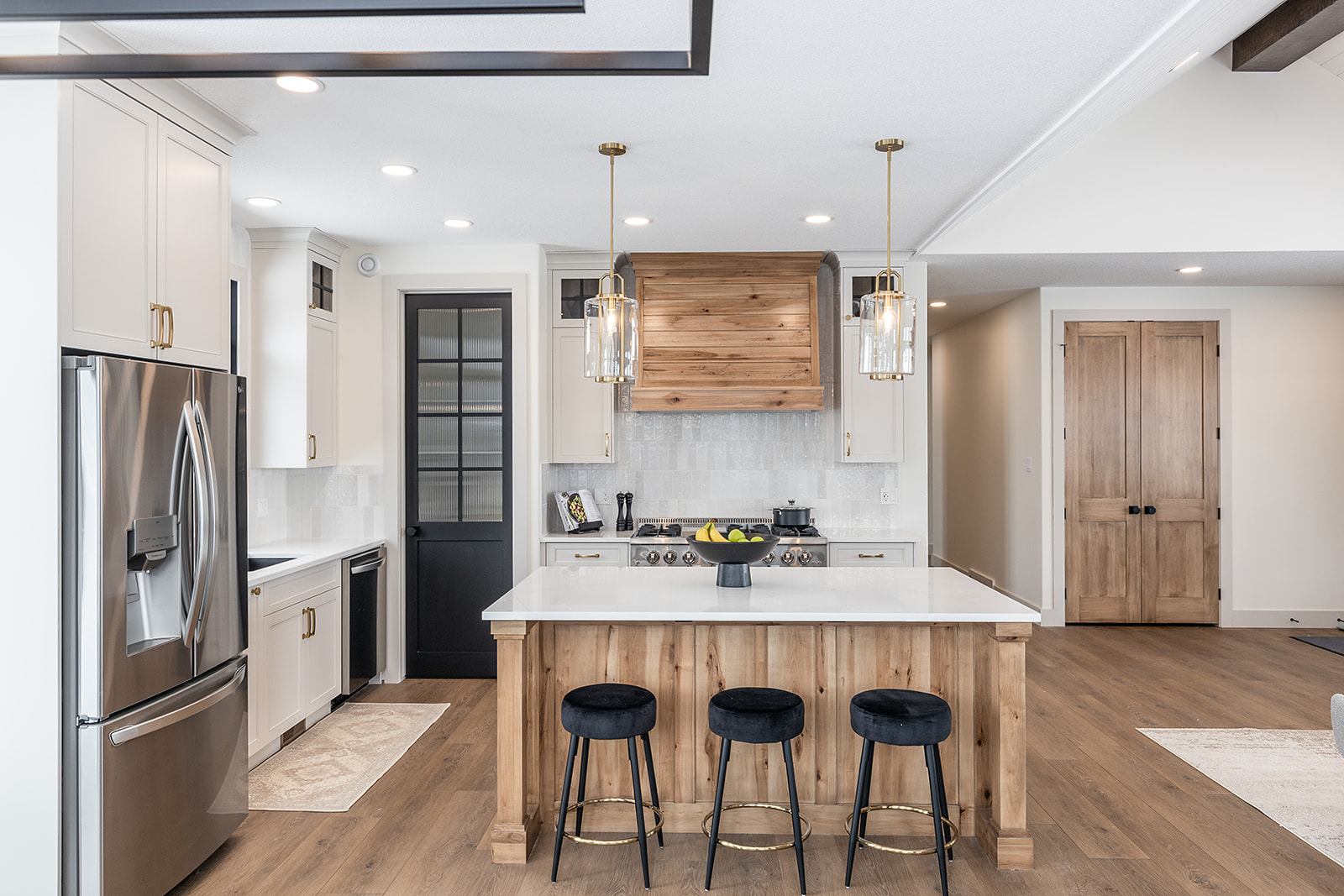 Modern kitchen with wood accents, featuring a central island with three black stools, stainless steel fridge, pendant lights, and light wood flooring.