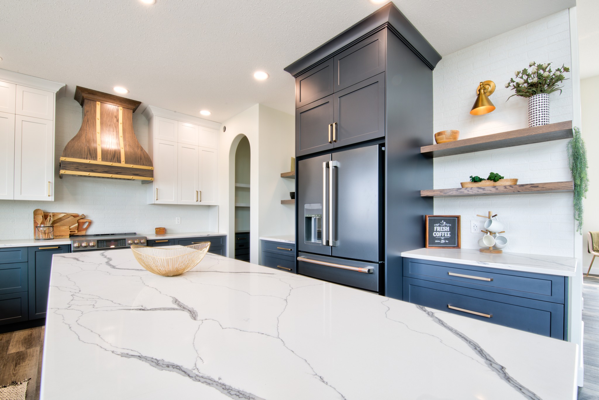 Modern kitchen with blue and white cabinets, large marble island, wood range hood, sleek black fridge, and floating shelves with plants and decor.