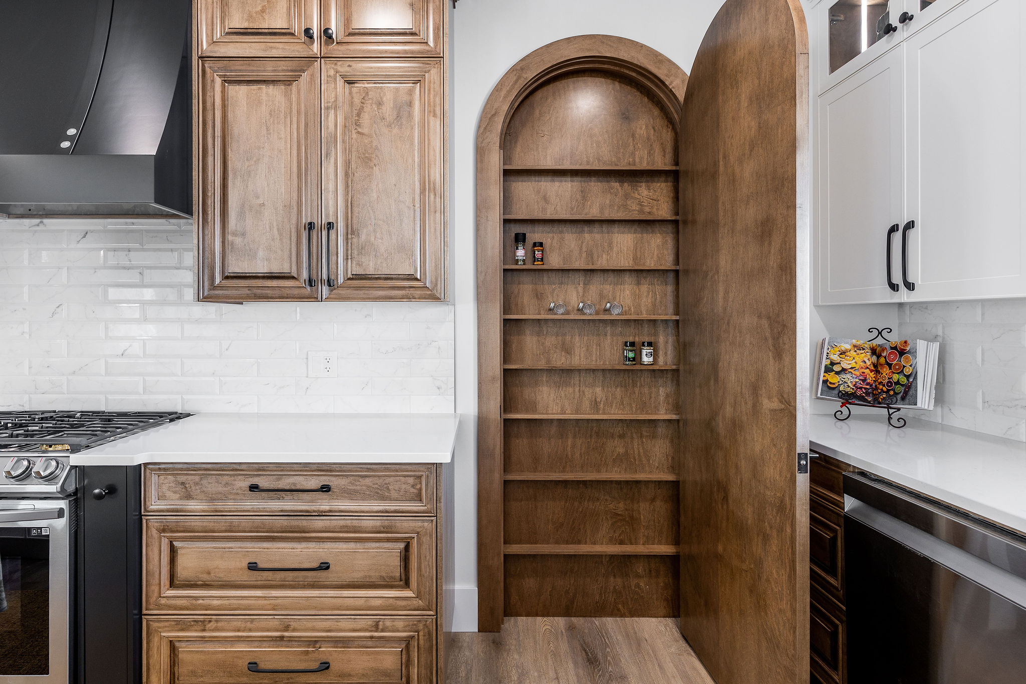 Kitchen with wooden cabinets and white countertops. A tall, arched, open pantry door reveals shelves with spices. Sleek, modern, and organized design.