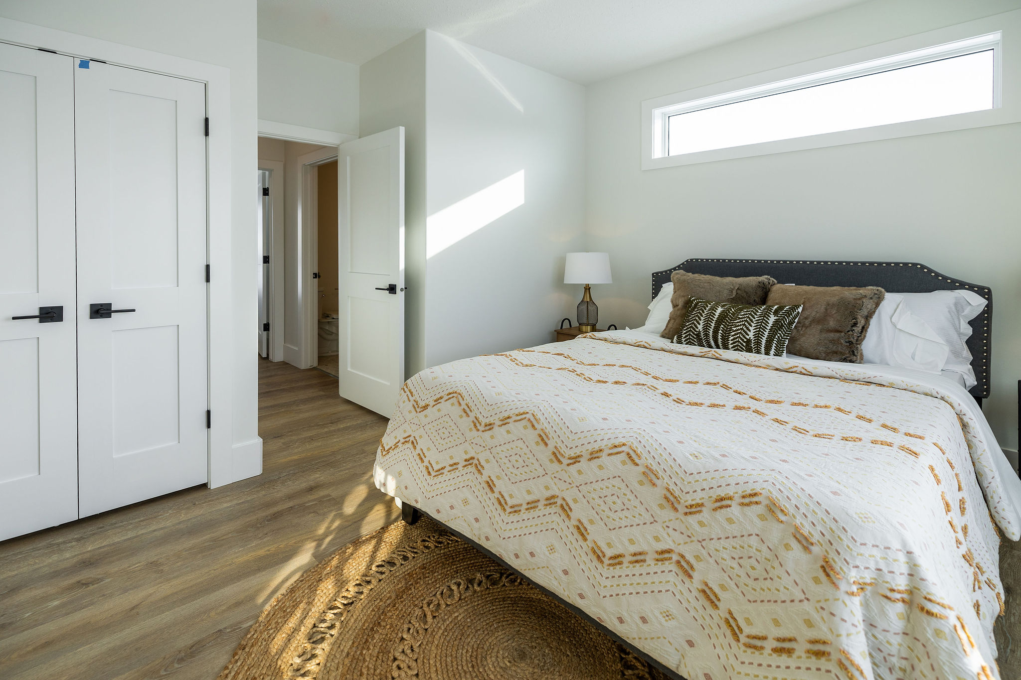 A bright, minimalist bedroom with a patterned bedspread, pillows, and bedside lamp. Wooden floor and sunlit walls create a cozy, inviting feel.