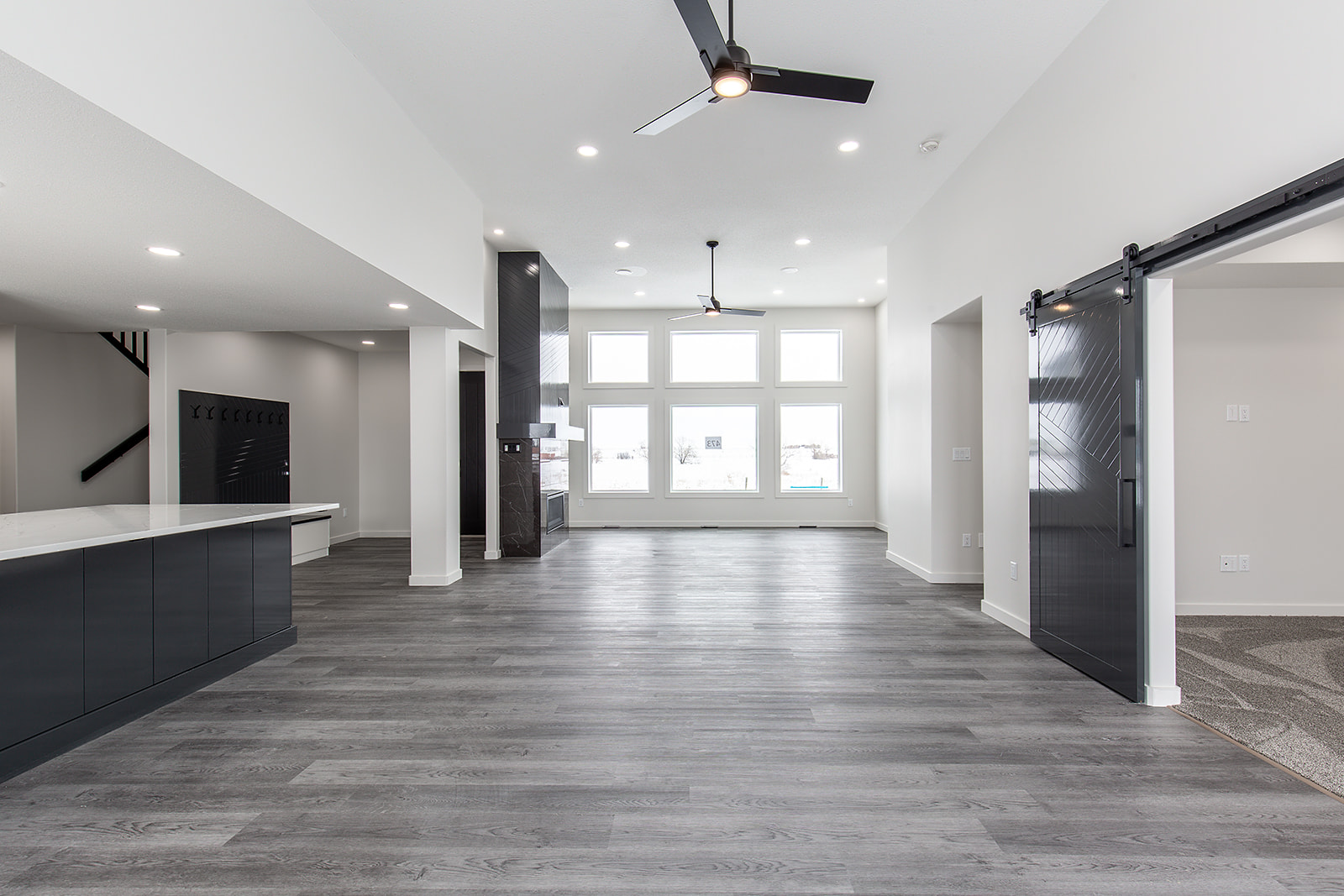 Spacious modern living area with gray wood flooring, large windows, white walls, ceiling fans, open kitchen, and a sliding barn door entry.