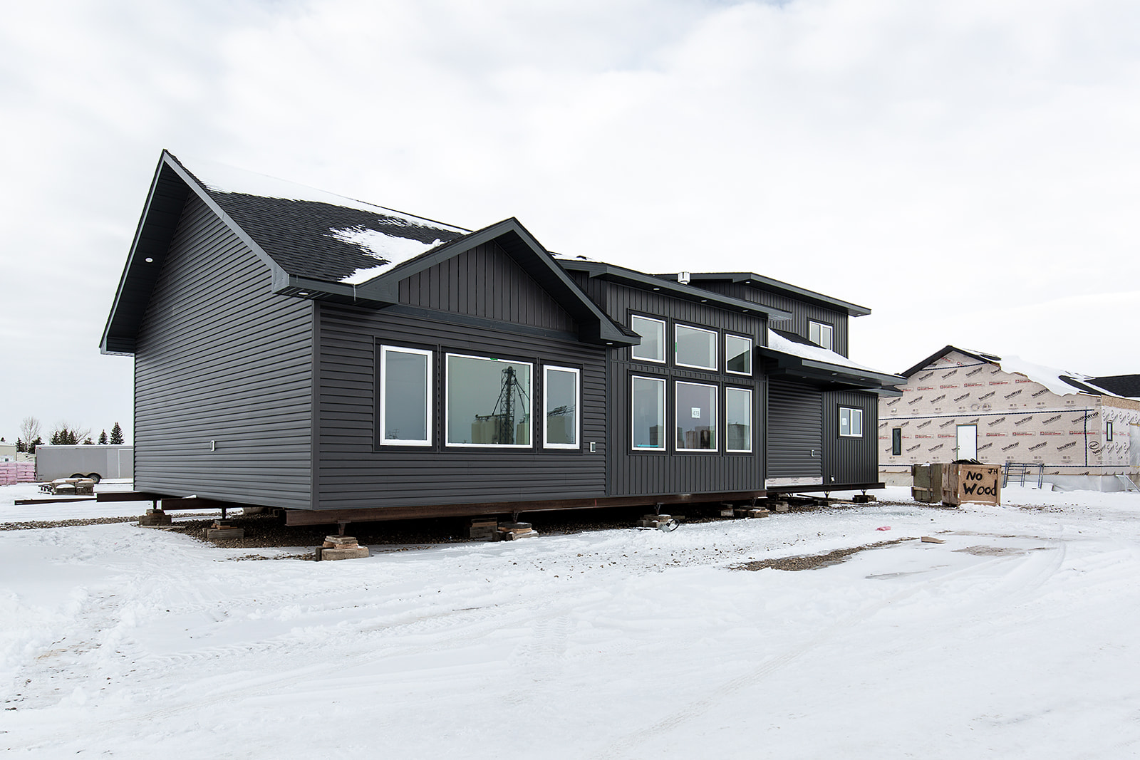 A gray modular home sits elevated on blocks in a snowy lot under a cloudy sky. Nearby, signs of construction and a "No Wood" sign are visible.