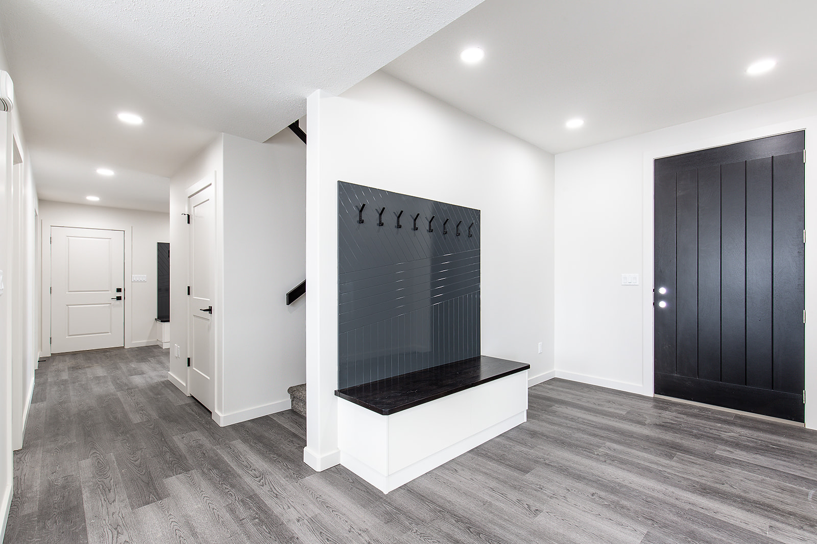 Modern, minimalist entryway with dark wood flooring. A black door and bench with coat hooks create a sleek look, enhanced by bright, recessed lighting.