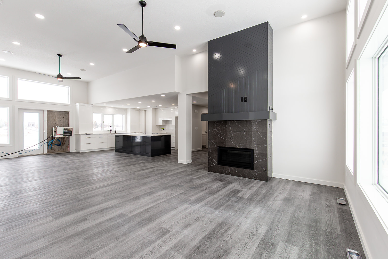Spacious modern living room with gray wood floors, large black fireplace, high ceilings, black ceiling fans, and abundant natural light from big windows.