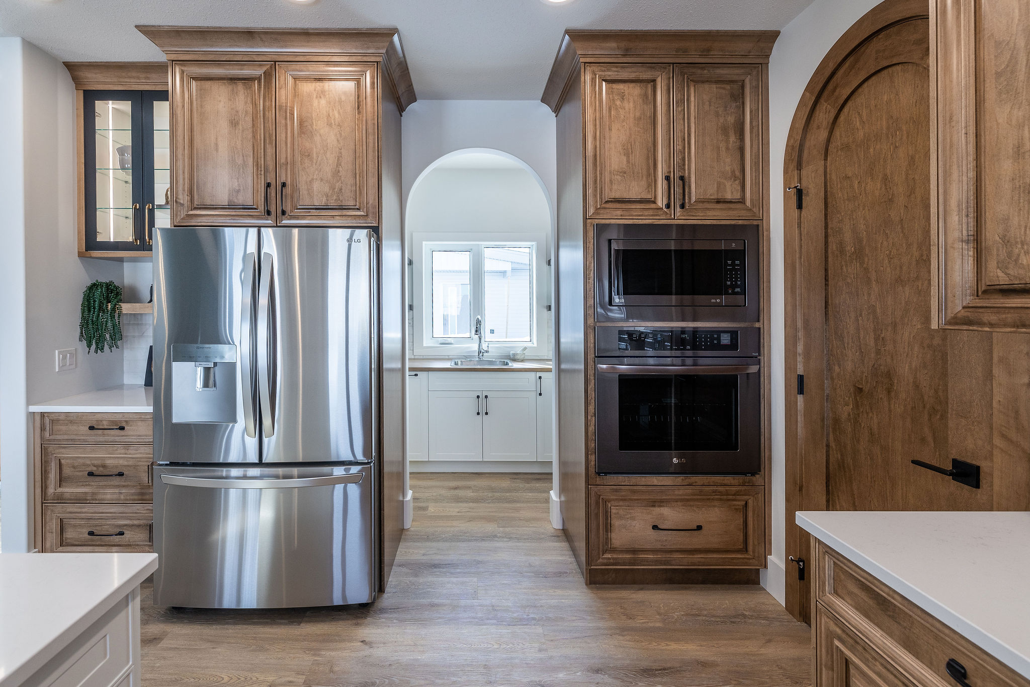 Modern kitchen with stainless steel fridge, wooden cabinets, arched doorway leading to a sink under a window, and built-in black ovens. Cozy and elegant.