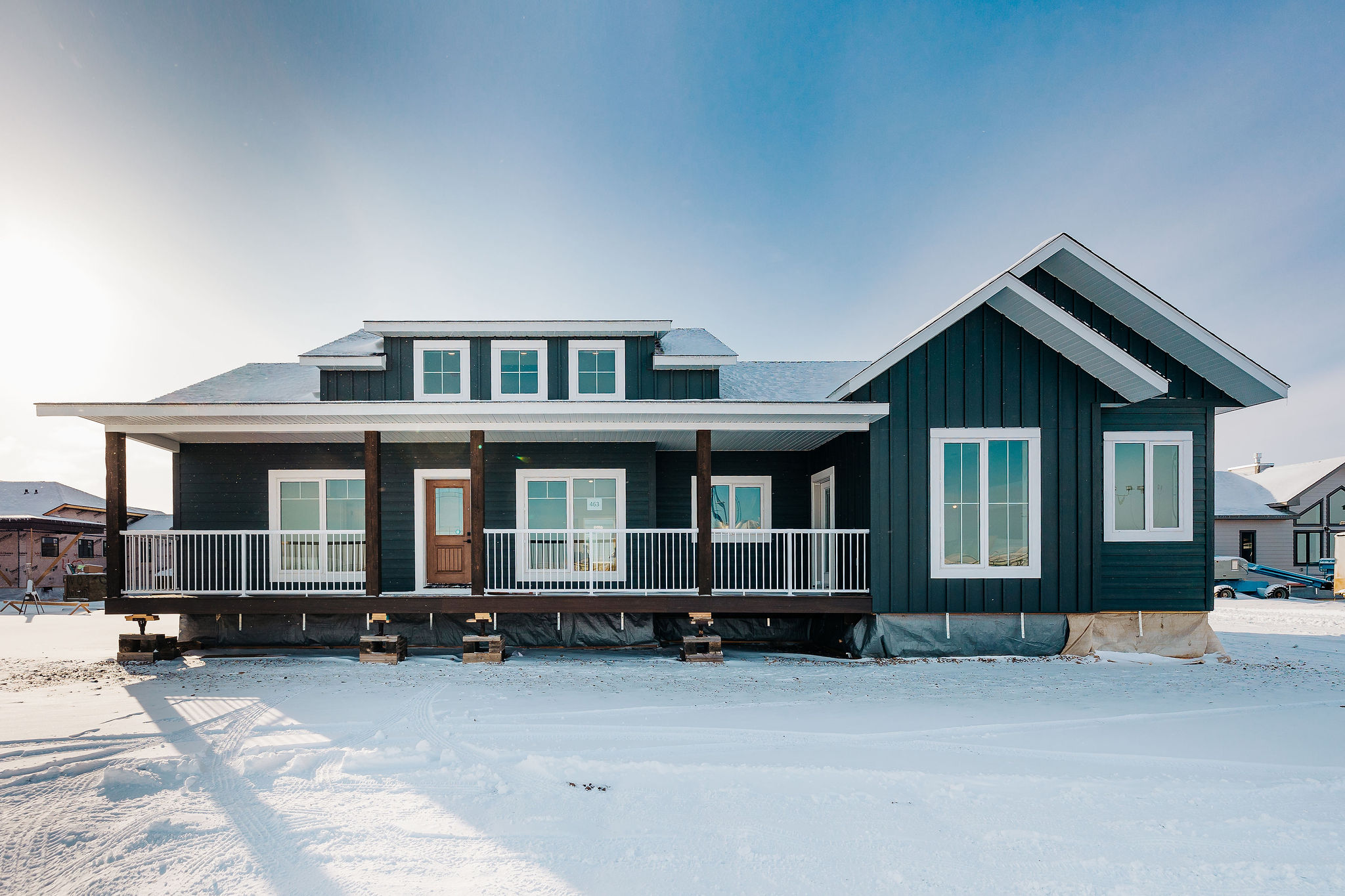 A dark teal, single-story house with white trim and a snow-covered roof sits on a snowy landscape. A wooden porch and railing add warmth to the scene.