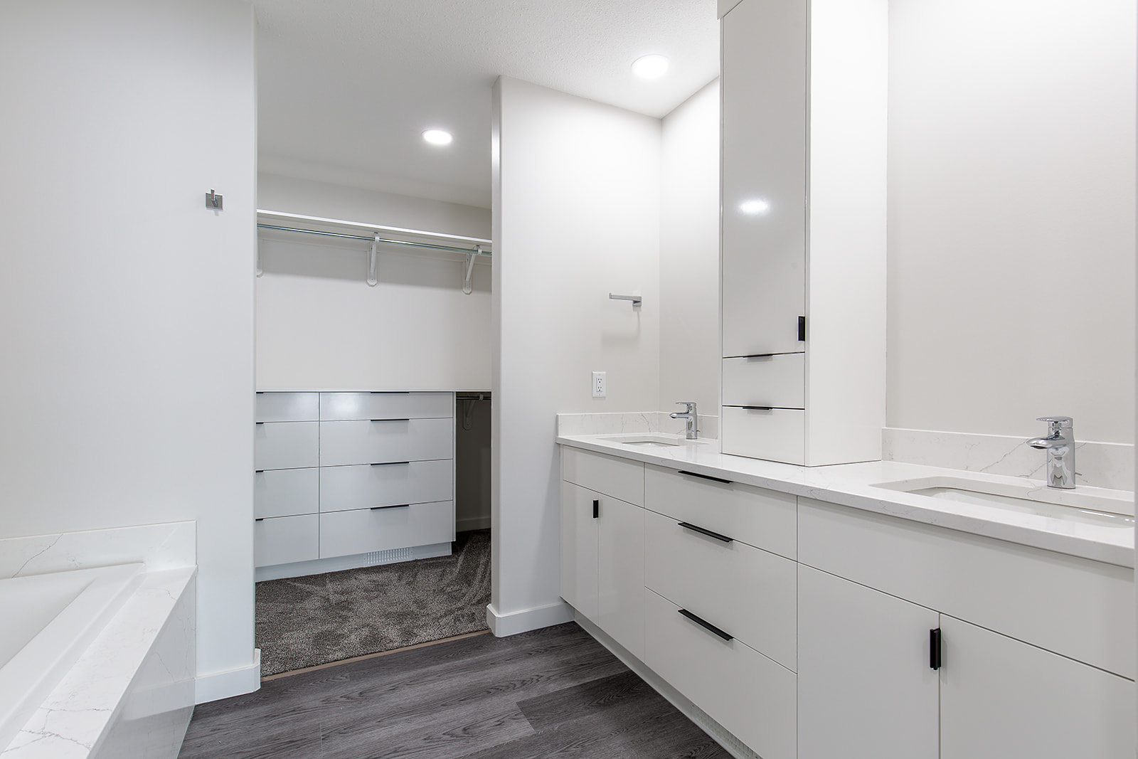 Sleek, modern bathroom with dual sinks, white cabinetry, and dark wood flooring. Bright lighting creates a clean, minimalist feel. Open closet space visible.