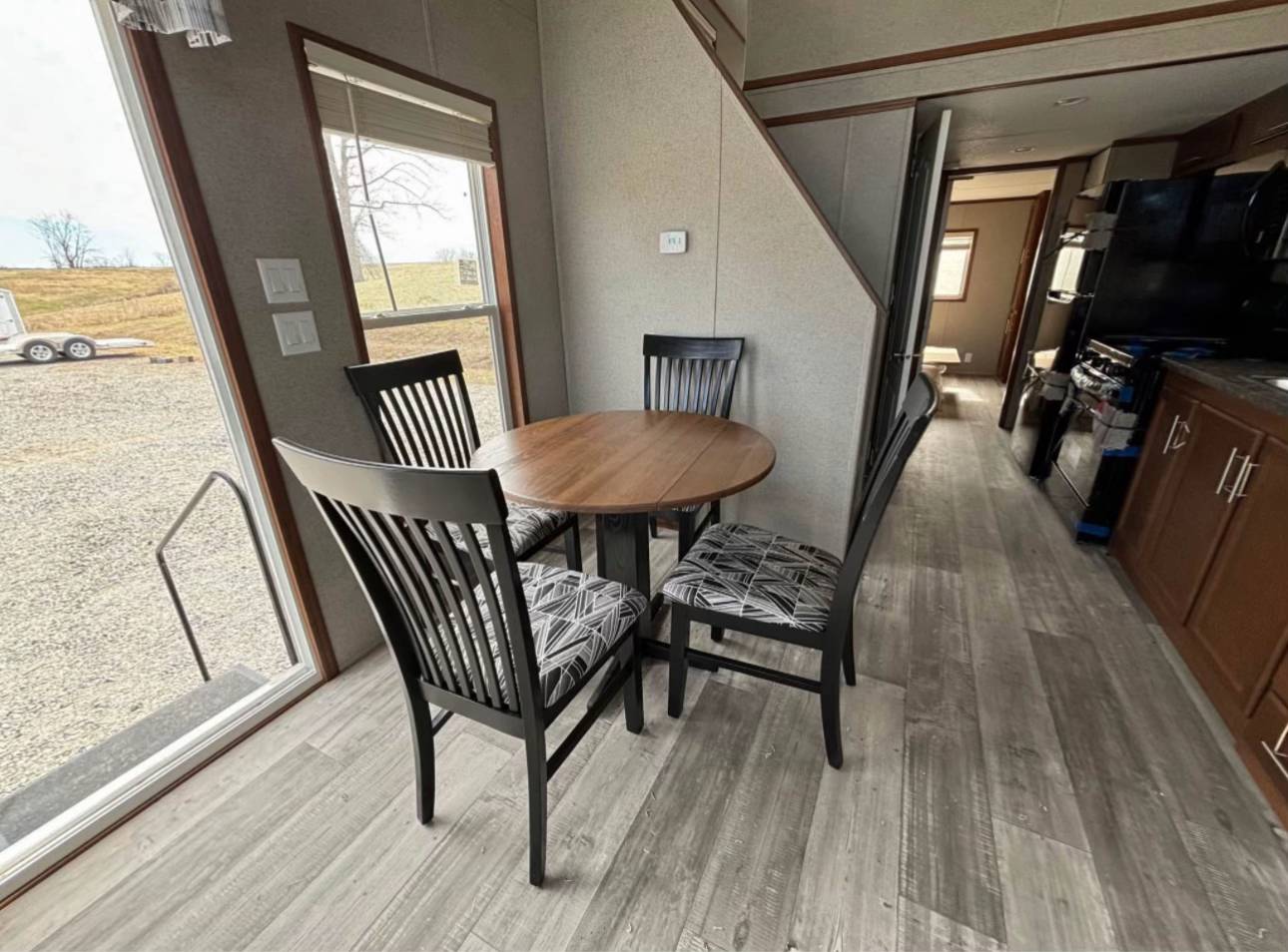 Cozy dining area with a round wooden table and four black chairs featuring patterned cushions, adjacent to a window overlooking a rural landscape.