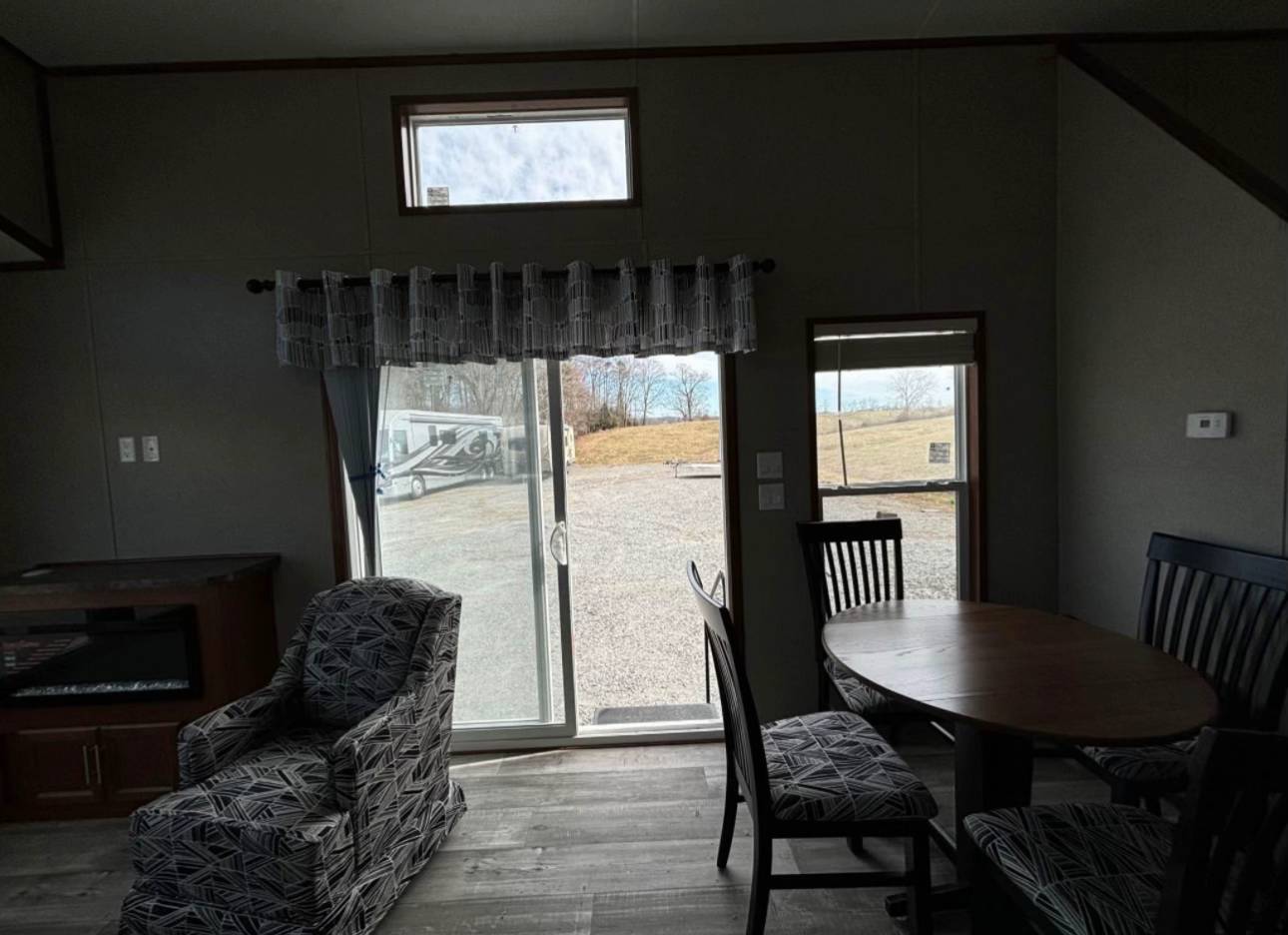Interior of a room with a round wooden table, chairs, patterned armchair, and a sliding glass door leading outside. Natural light enters, creating a cozy feel.