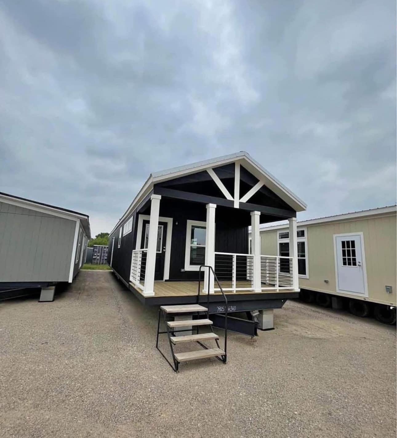 A modern, dark tiny home with white trim and a small porch stands on gravel under a cloudy sky. It is elevated with steps leading to the entrance.
