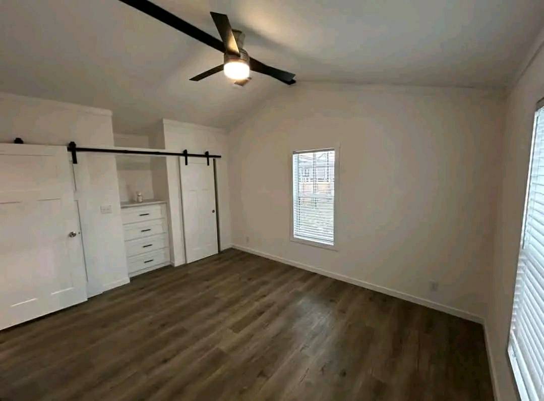 A spacious bedroom with light wood flooring and white walls. It features a ceiling fan, two windows with blinds, and sliding barn doors. The room feels modern and airy.