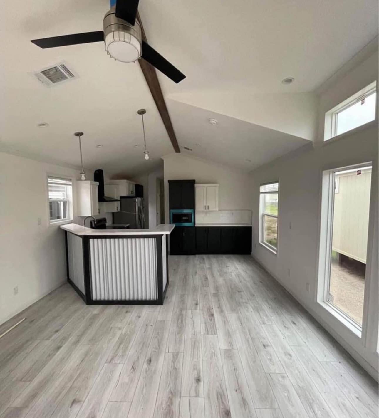 Modern kitchen with light wood flooring, white walls, and large windows. Features a black and white island, sleek cabinetry, and ceiling fan. Bright and airy.