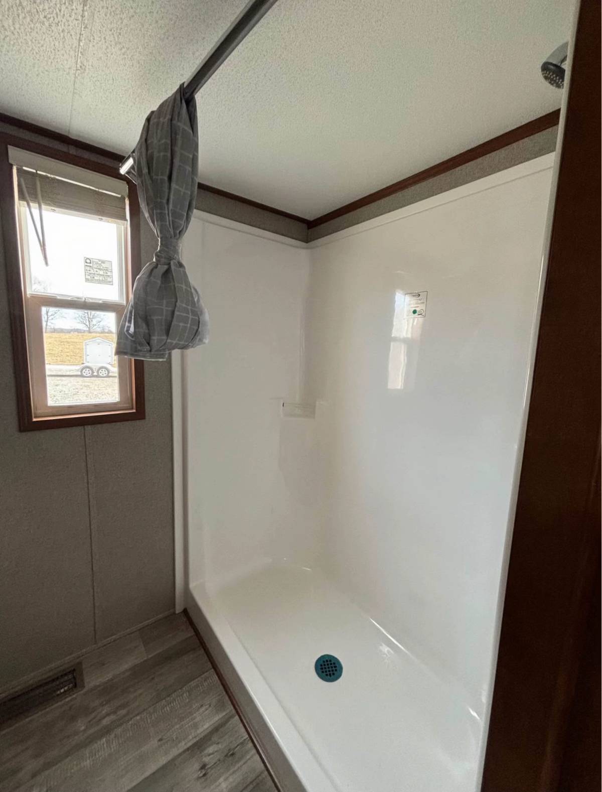 Shower stall with white walls and a plaid gray curtain tied to the side, wooden trim, and large window letting in natural light, creating a fresh atmosphere.