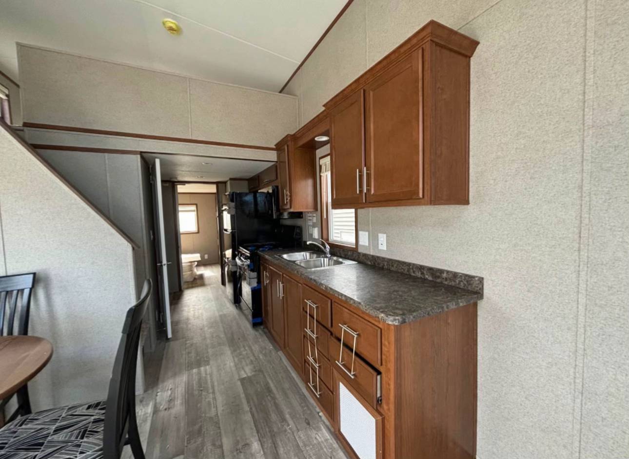 Compact kitchen in a mobile home, featuring wooden cabinets, a dark countertop, stainless steel sink, and black appliances. Minimalist and functional.