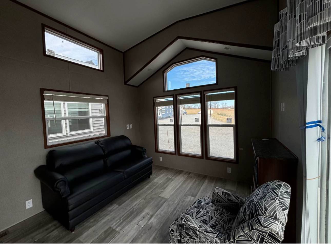 Modern living room with high ceilings and large windows showcasing an exterior view. Features a black sofa, patterned armchair, and wooden floors. Bright, airy ambiance.