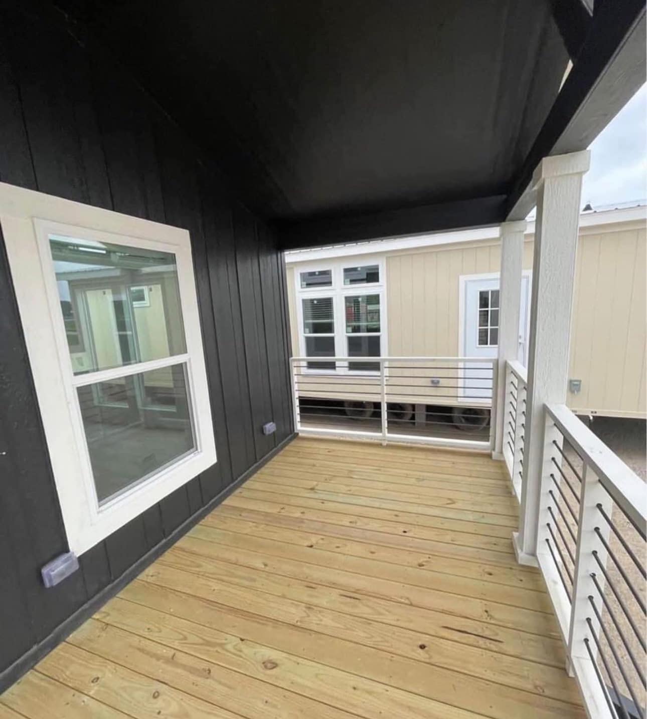 Covered porch with wooden decking, black siding, and white railing. Adjacent structure with beige exterior and large windows visible. Calm atmosphere.