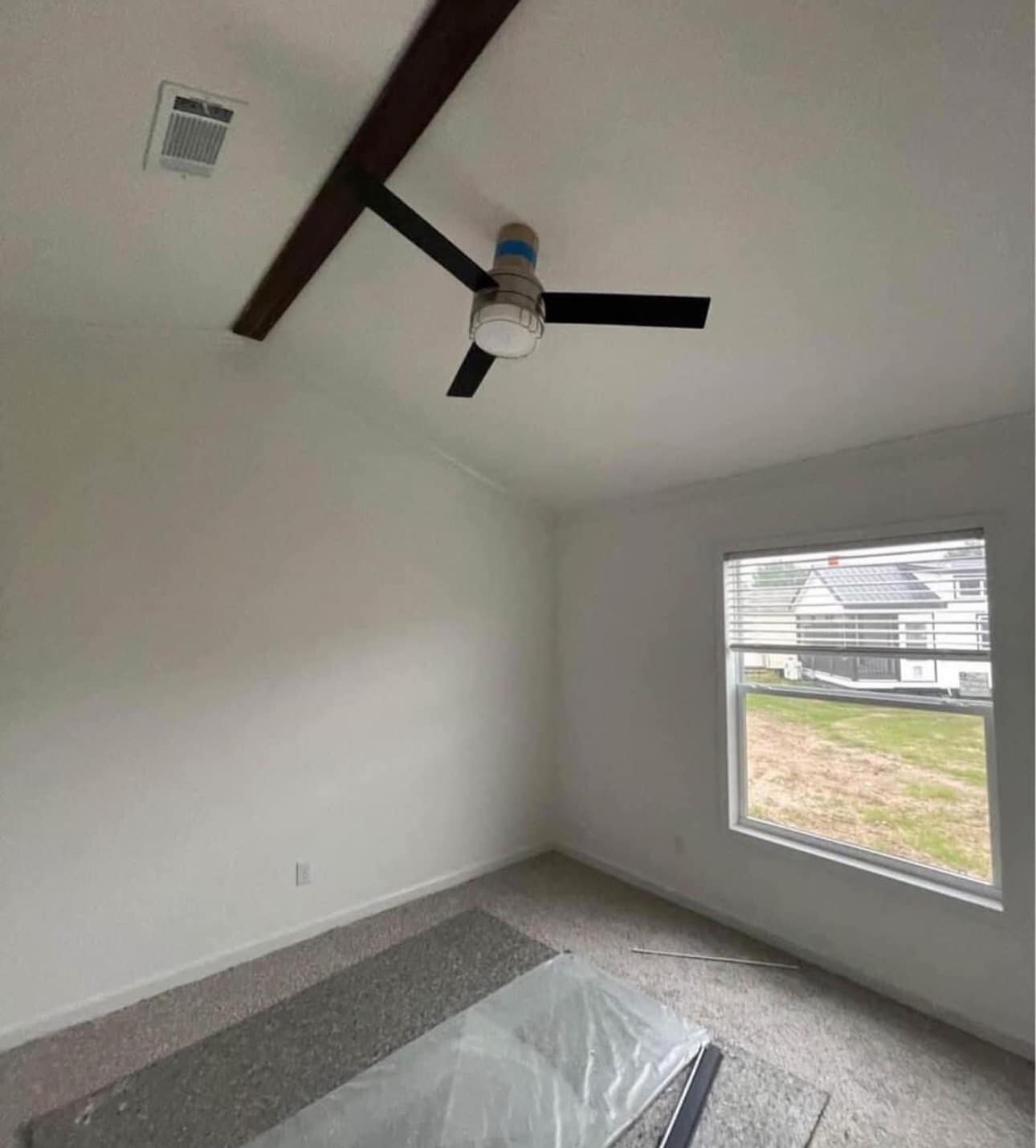 A minimalist room with white walls, brown ceiling beam, and dark ceiling fan. Sunlit window showing houses outside. Carpeted floor with protective wrap.