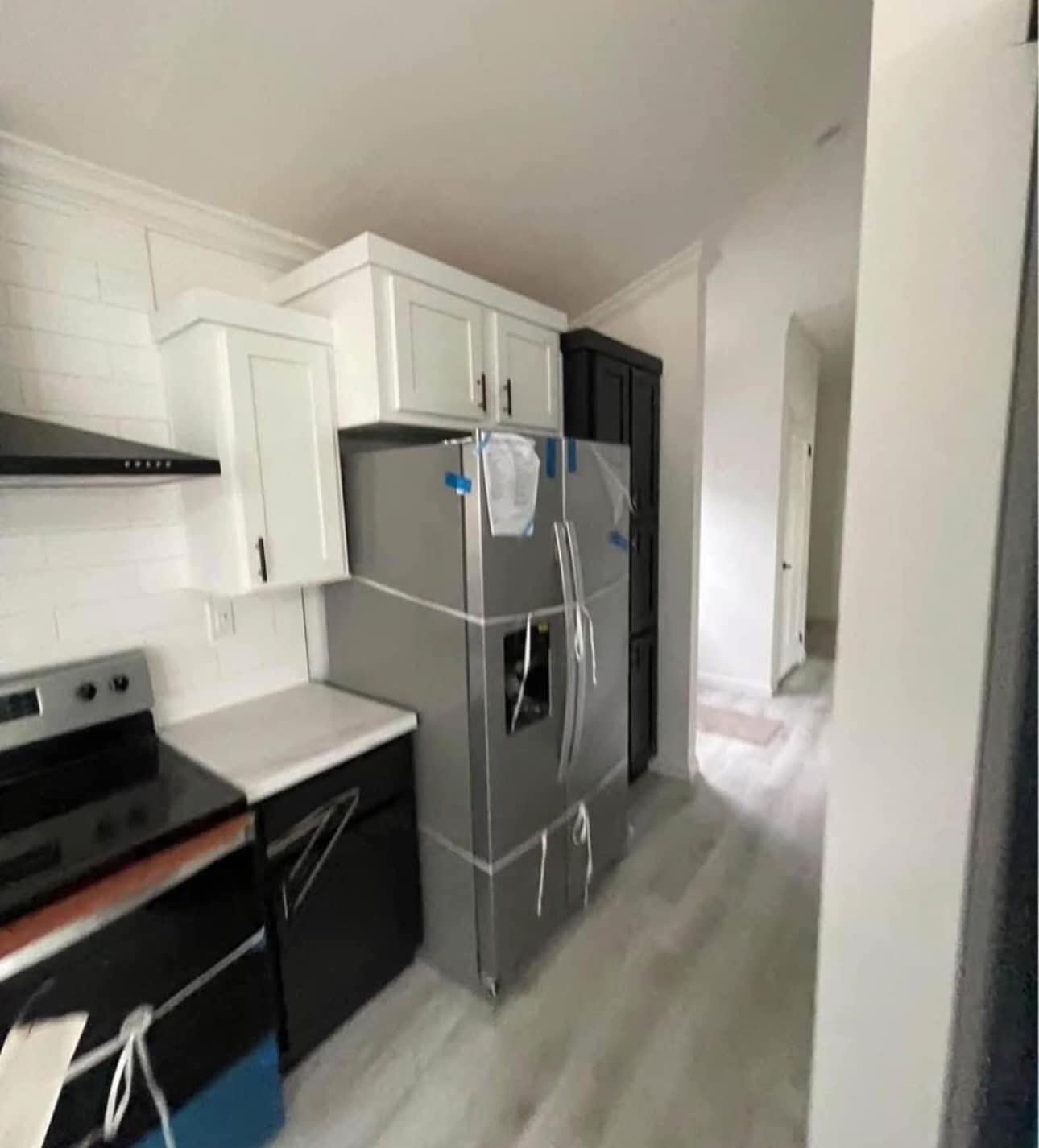 A modern kitchen with white cabinets, a stainless steel fridge, and stove. Appliances are wrapped, suggesting recent installation. The hallway leads to another room.