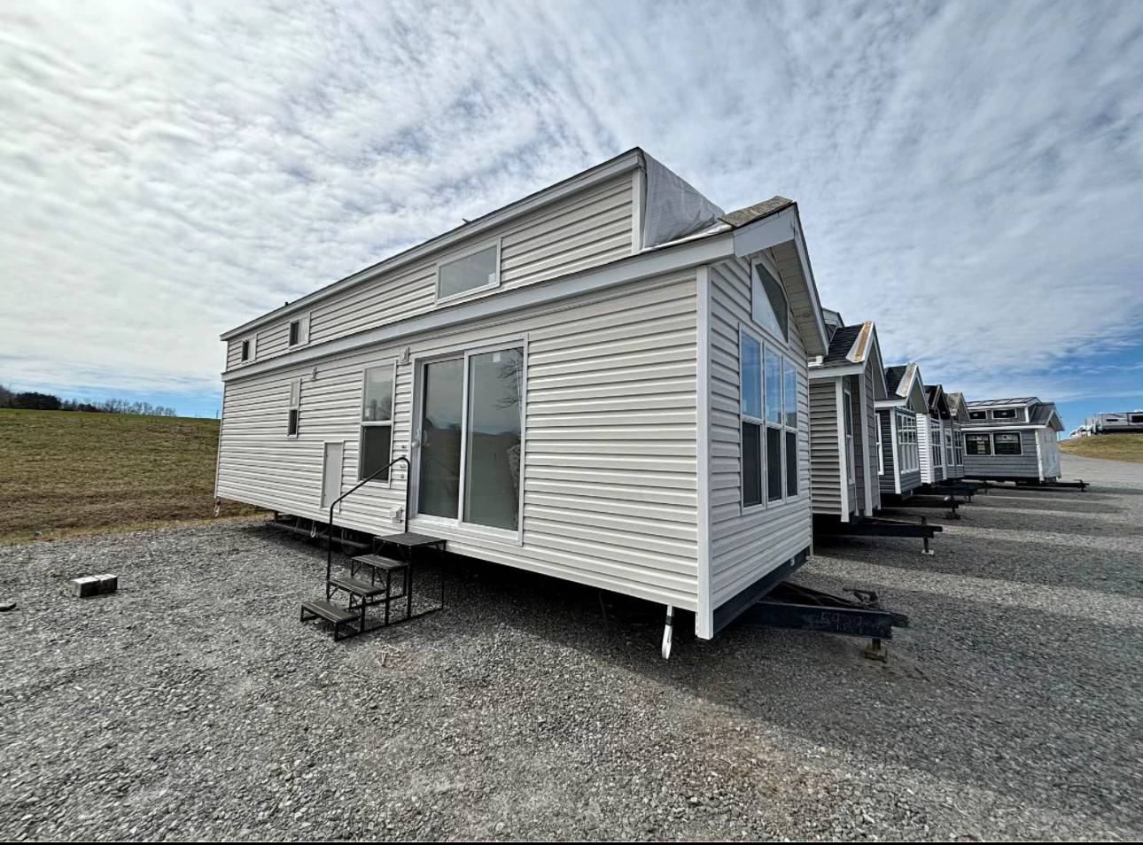Row of white, modern tiny houses with large windows and steps, set on a gravel lot under a partly cloudy sky, evoking a minimalist, serene atmosphere.