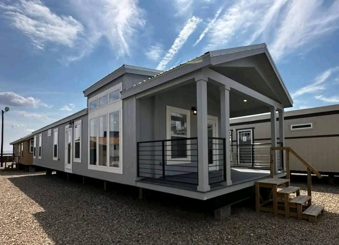A gray and white mobile home parked on a gravel surface, surrounded by a clear sky.