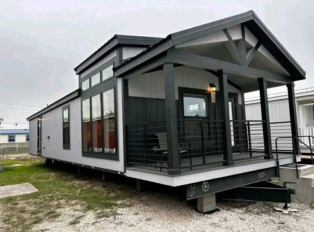 Modern tiny house with grey siding, large windows, and a covered porch. It sits elevated on a gravel lot, exuding a cozy, contemporary vibe.