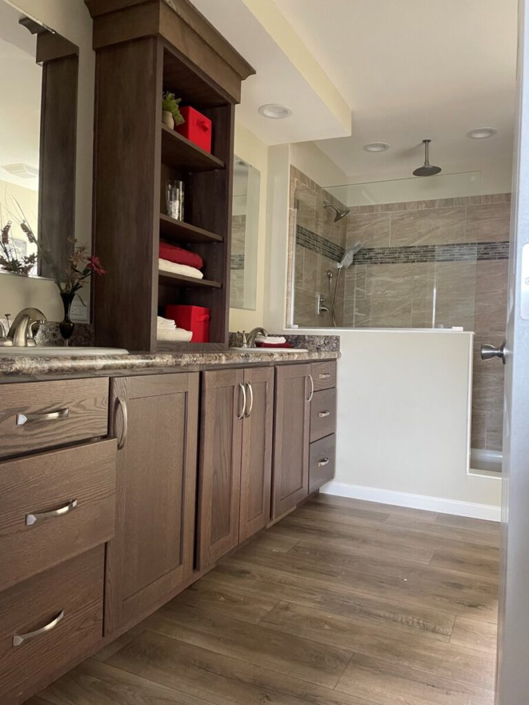 Modern bathroom with dark wood cabinets, open shelving displaying red and white towels, granite countertop with dual sinks, and a glass-enclosed shower.