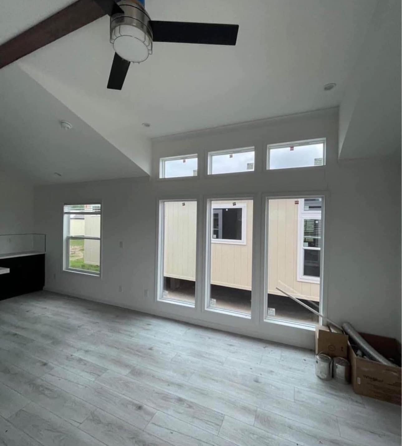Spacious, unfinished room with light wood flooring, large rectangular windows, and ceiling fan. Outside, cream building and boxes in corner suggest construction.