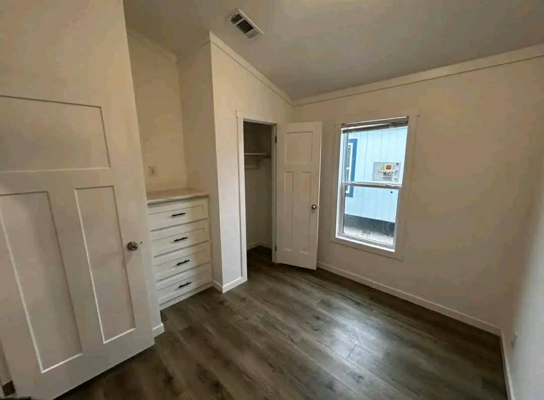 A small, empty room with white walls, wood flooring, a window, and open closet on the right. Built-in drawers are on the left, under soft lighting.