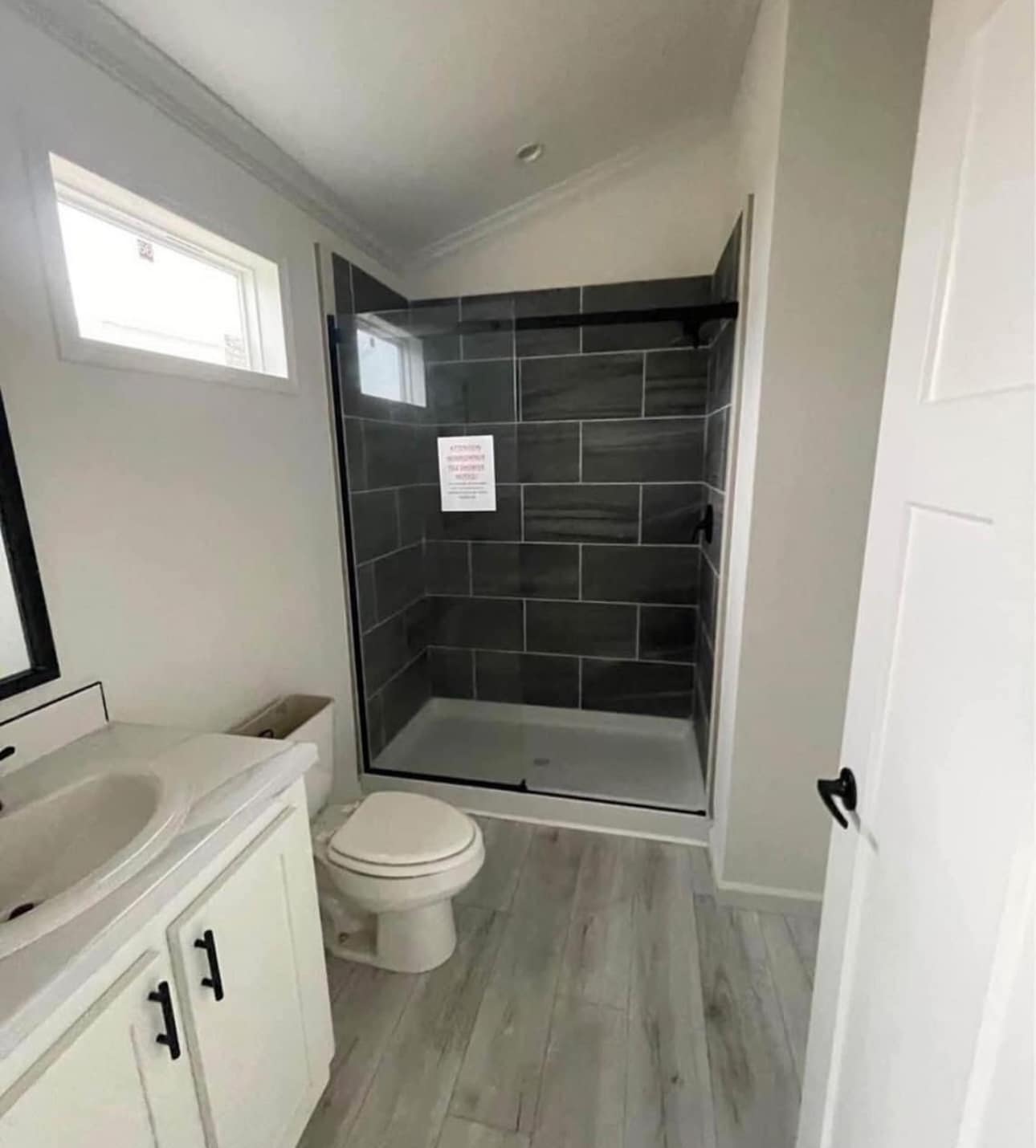 Small modern bathroom with gray-tiled shower, glass door, and a white toilet. Light wood-style flooring and a white vanity create a clean look.