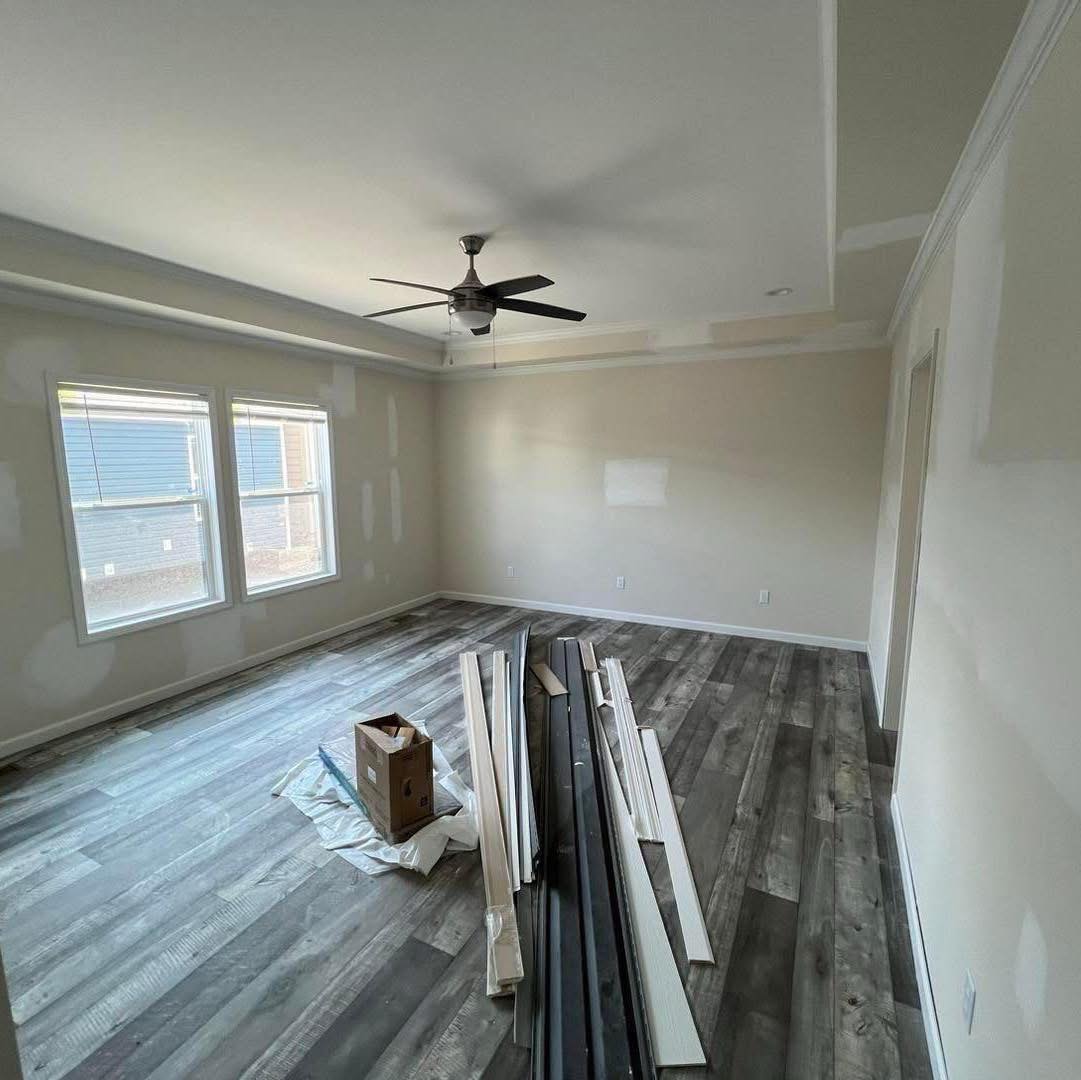 A spacious, unfinished room with gray wood floors, beige walls, a ceiling fan, and two windows. Lumber is stacked on the floor, suggesting ongoing renovations.