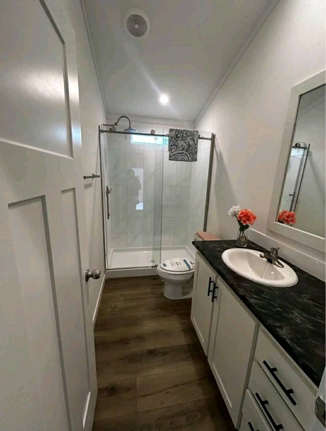 Modern bathroom with wood flooring, a glass shower, and white walls. A black countertop with a sink and pink flowers add a touch of color.