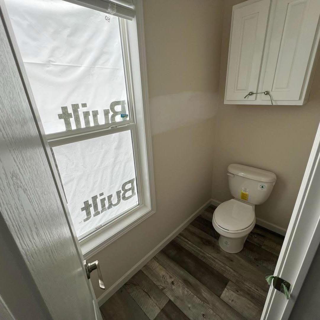 A small, narrow bathroom with light wood flooring, a white toilet, a wall cabinet, and a window with a "Built" sign partially visible outside.