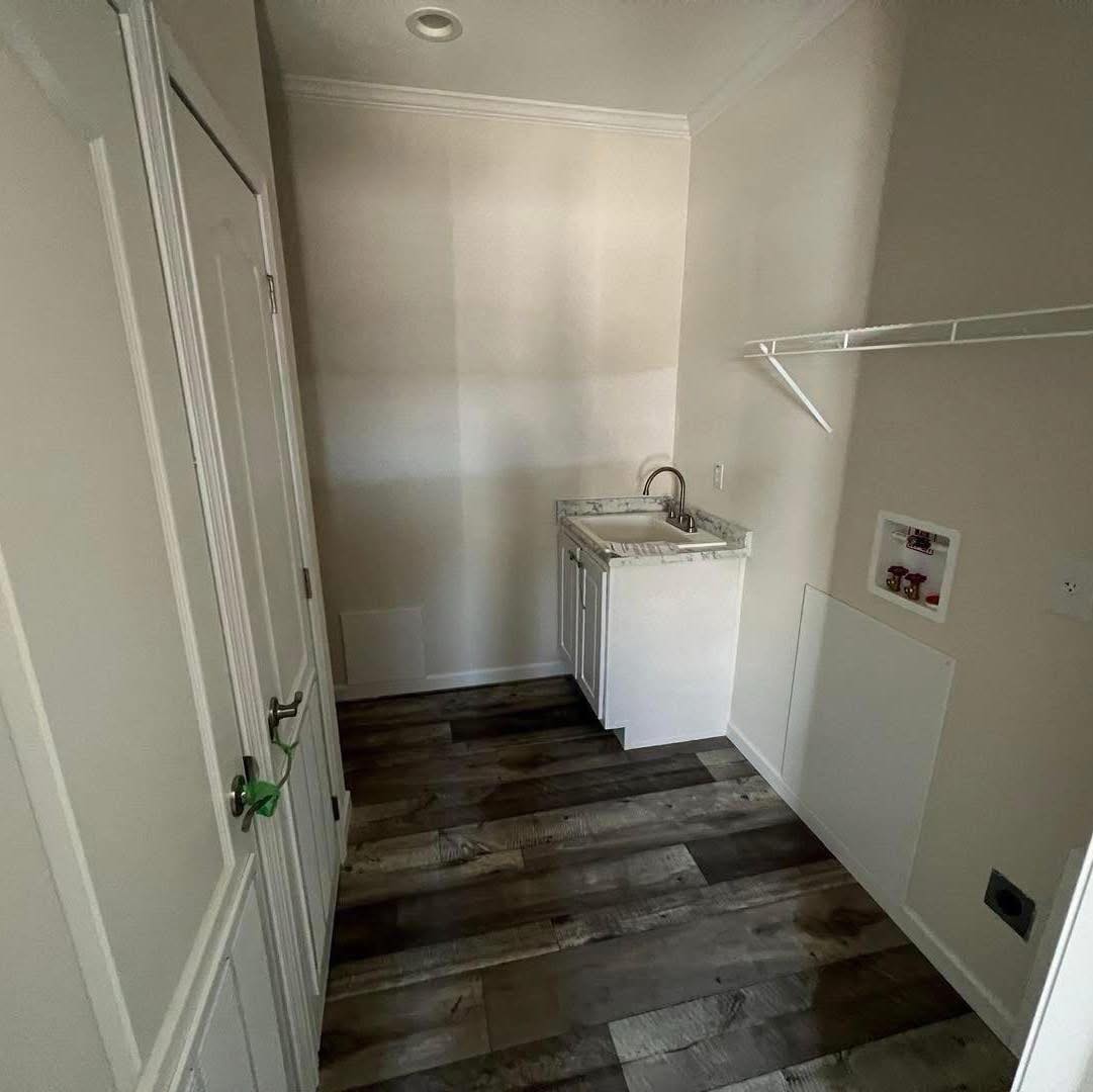 Empty laundry room with light beige walls, a small white sink with a marble counter, wooden floor, shelf, and plumbing connections. Simple and unused.