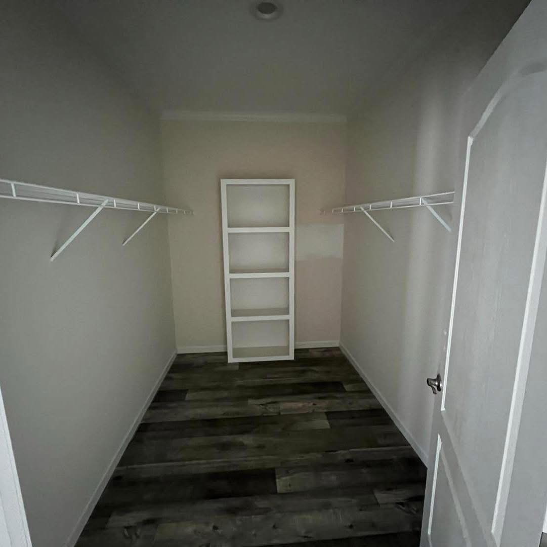 A narrow, empty walk-in closet with white walls and dark wood flooring. Two long white wire shelves run along both sides, with a shelf unit at the end.