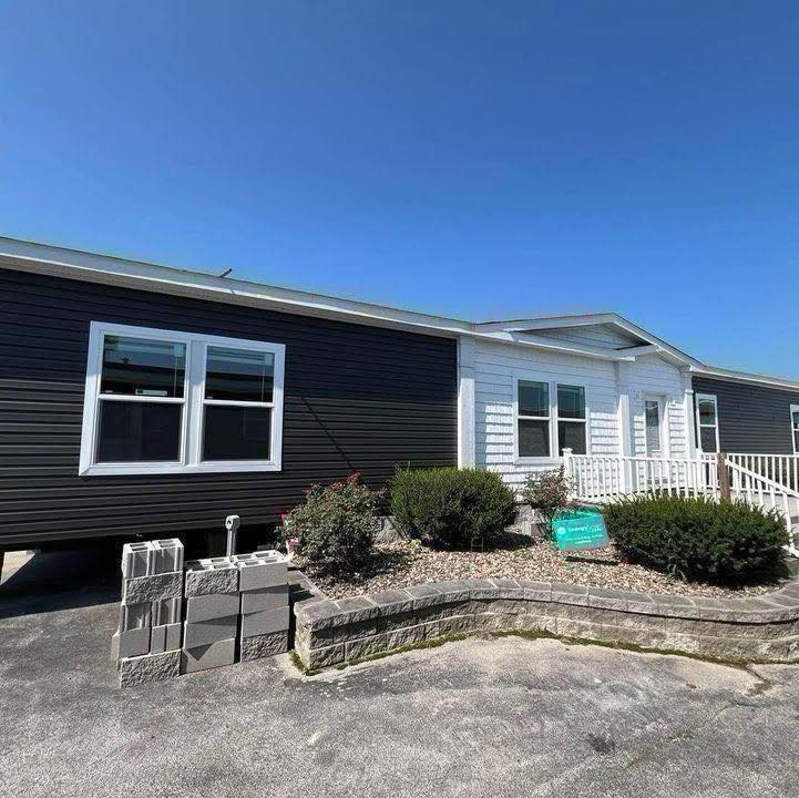 Single-story modular home with dark siding and white trim, fronted by shrubs and a stone-lined garden. Bright blue sky overhead, creating a welcoming feel.