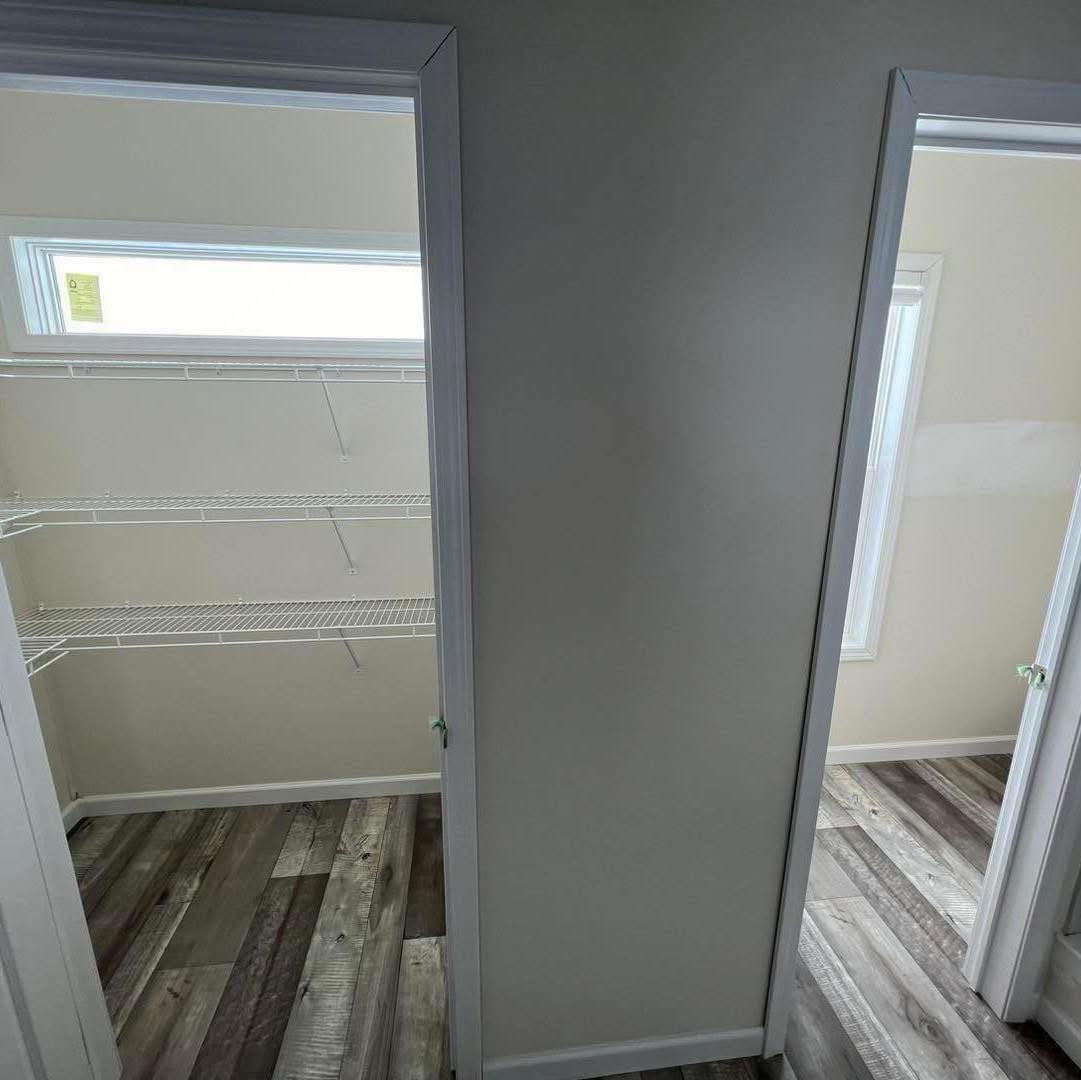 A narrow hallway features two open doors. On the left, a closet with white wire shelves and a narrow window. The floor has a rustic wood pattern.