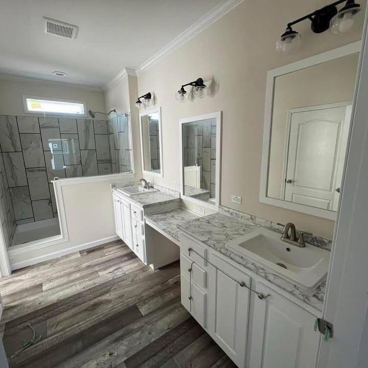 Bright bathroom with dual sinks, marble countertops, white cabinets, and a large tiled shower. Warm wood floor and sleek, modern light fixtures.