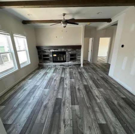 Spacious living room with light gray wood flooring, a brick fireplace with a dark wooden mantel, and large windows. Ceiling fan and exposed beam add warmth.