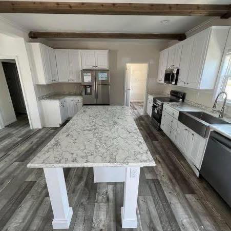Spacious kitchen with white cabinets, stainless steel appliances, and a large marble island. Wooden beams and gray flooring add warmth. Bright, modern ambiance.