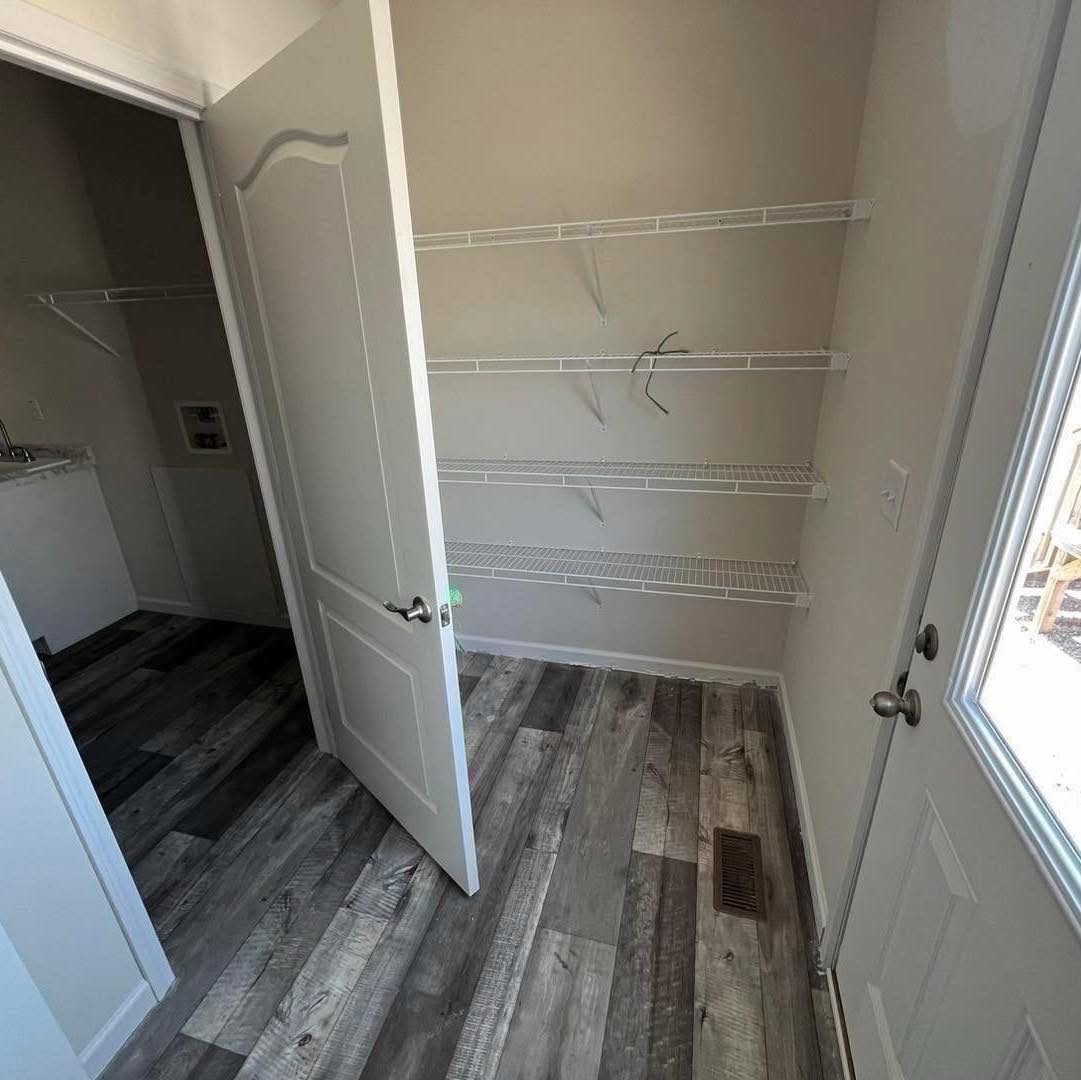 A small room with light gray walls and wooden flooring. It features white wire shelves on the right wall and an open door leading to another room.