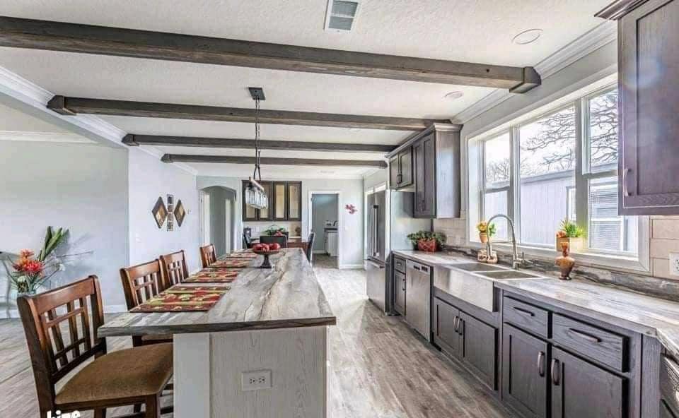 Spacious, modern kitchen with rustic wooden beams, a long island with chairs, dark cabinets, stainless steel appliances, and large windows.