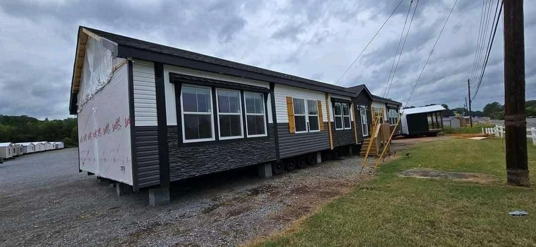 A modular home with dark gray siding and multiple windows is set on a gravel lot. Wooden stairs lead to the entrance. Overcast sky above.