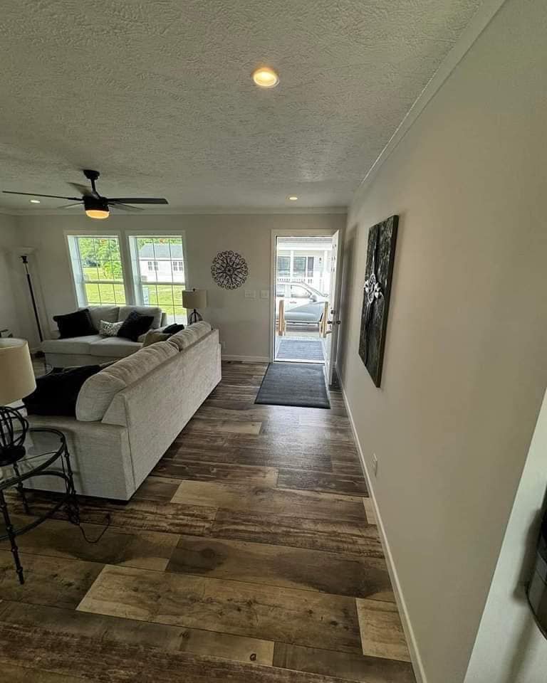 Spacious living room with light gray walls, wooden flooring, and a beige sectional sofa. Large windows and an open door let in natural light. A ceiling fan and wall art add modern elegance.